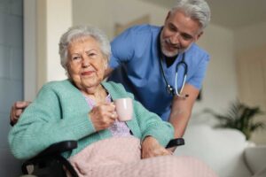Two older adults and a caregiver seated together at a sunny dining table, sharing conversation and gentle smiles.